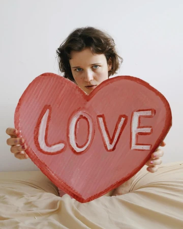 A young woman with dark hair in bed holds a large cardboard heart with the word 'Love' painted on it in a brightly lit, indoor setting.