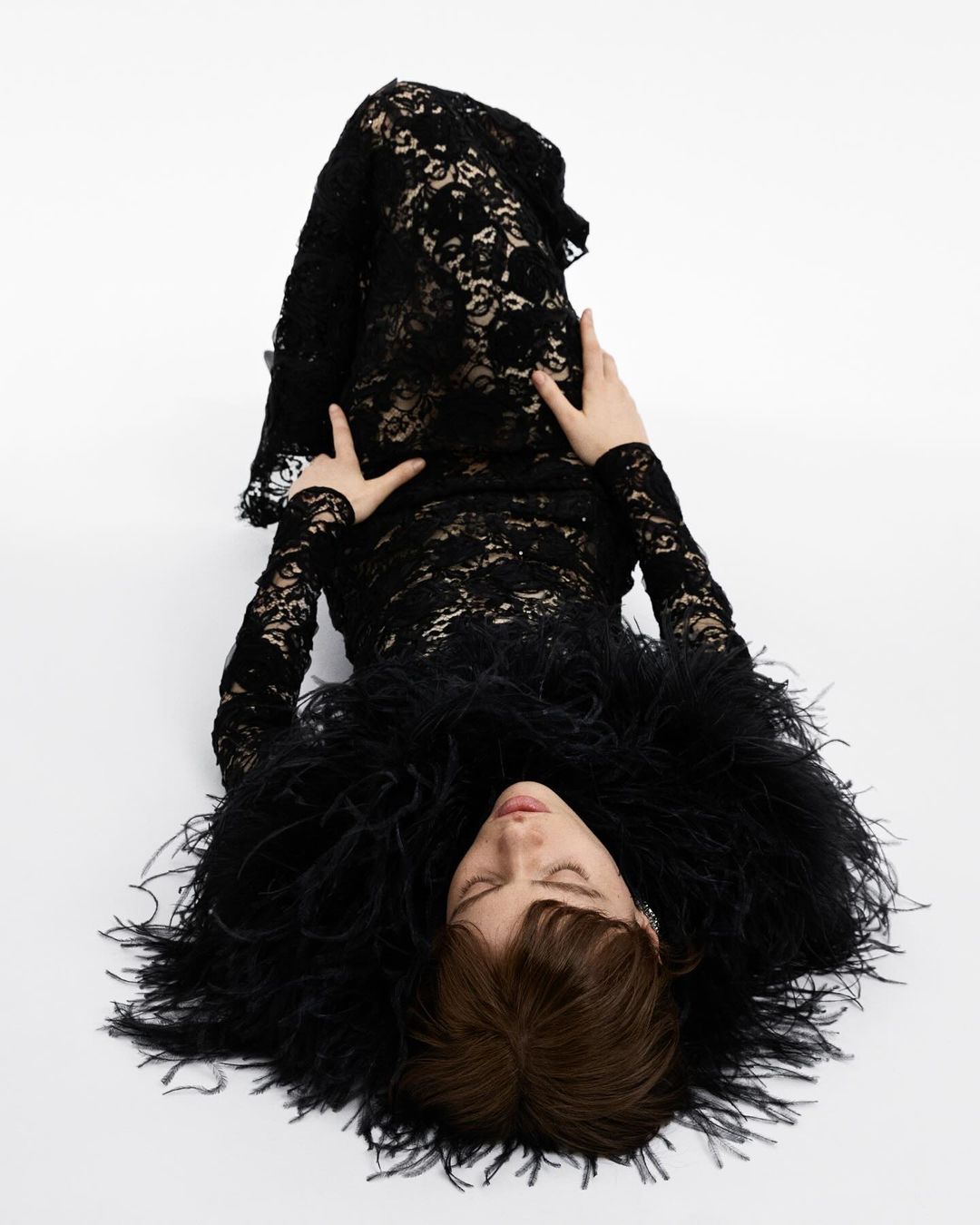 Woman in black lace dress with feather boa lying on white backdrop in a fashion shot. The image focuses on the model and her attire.
