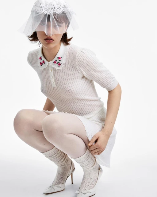 A slender woman poses in a white, short-sleeved dress with a floral-detailed collar and high heels against a stark white backdrop.