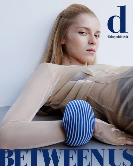 Portrait of a fair-skinned blonde woman in a sheer top, lying down next to a blue and white striped object, with the text BETWEEN US.