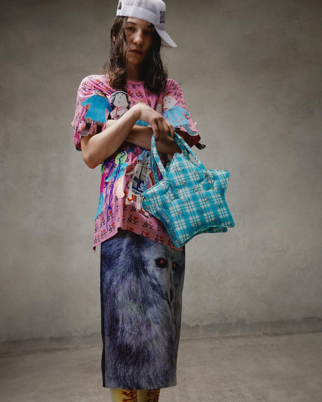 Adult female posing in a studio with pink graphic tee, wolf-print skirt, white baseball cap, and star-shaped handbag. Fashion editorial shot.