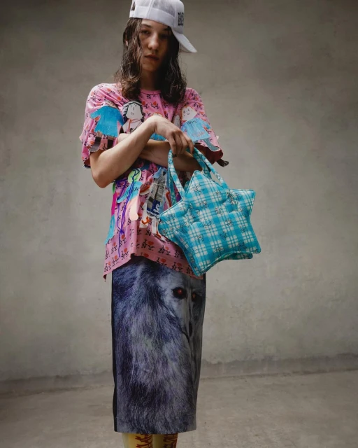 Adult female posing in a studio with pink graphic tee, wolf-print skirt, white baseball cap, and star-shaped handbag. Fashion editorial shot.