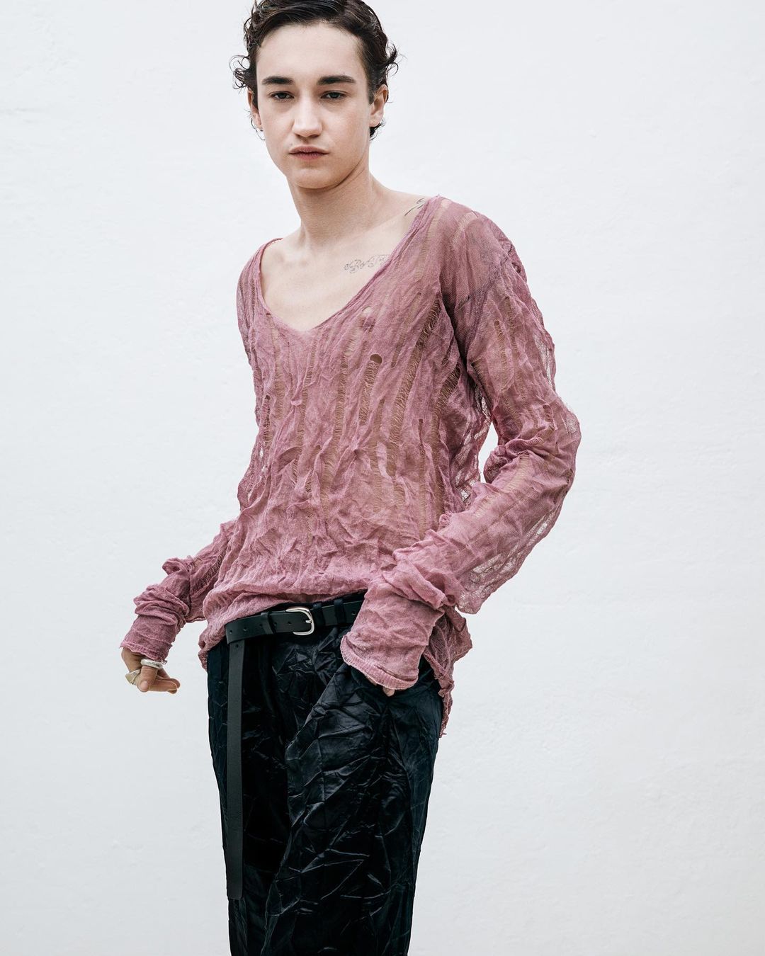 A person with dark hair stands in front of a neutral backdrop, wearing a textured pink V-neck long-sleeve shirt and dark, shiny trousers.