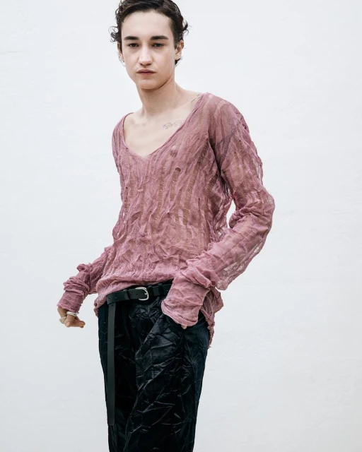 A person with dark hair stands in front of a neutral backdrop, wearing a textured pink V-neck long-sleeve shirt and dark, shiny trousers.