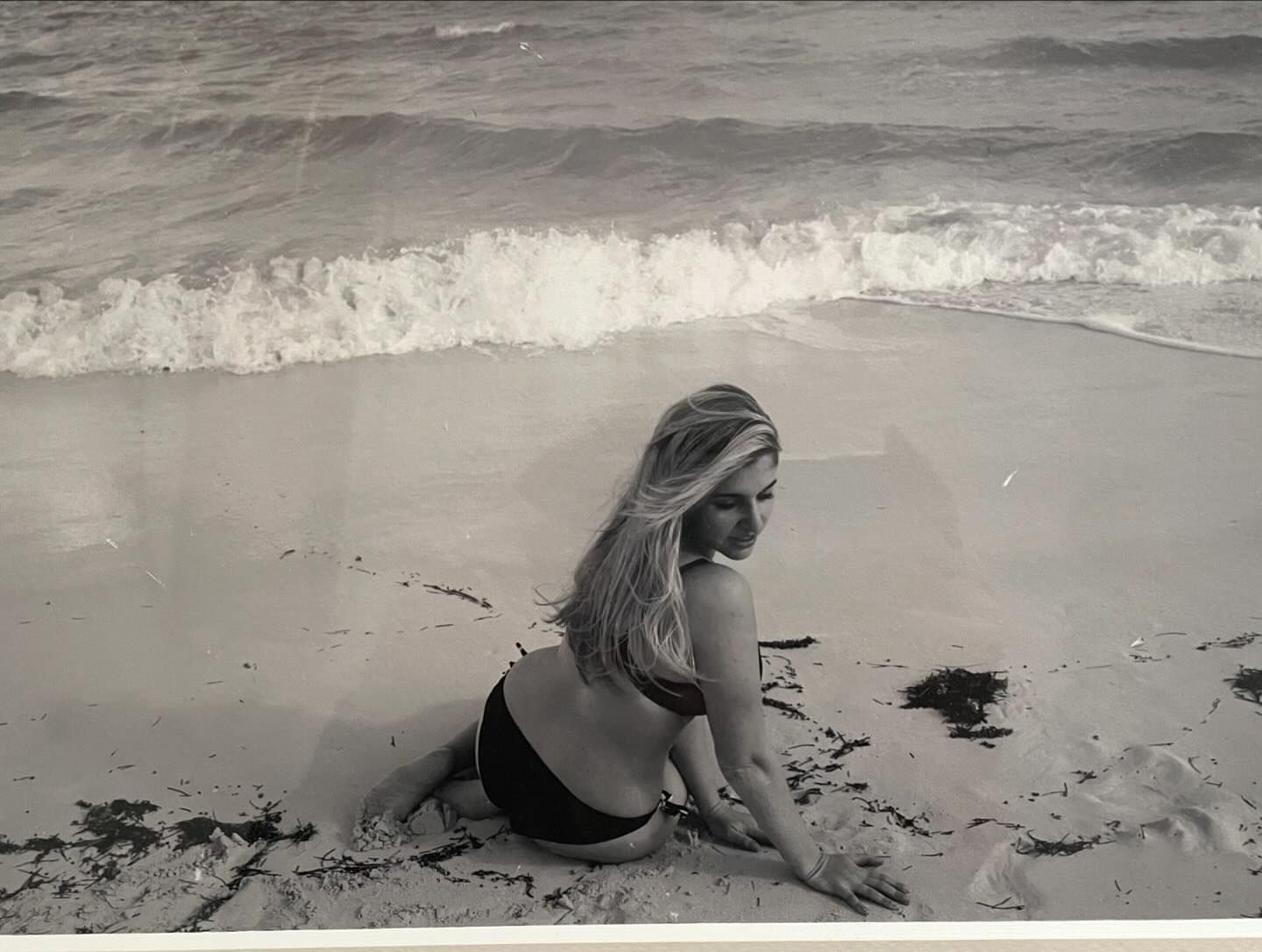 Monochrome photo of a woman in swimwear sitting on a sandy beach, with ocean waves in the background, captured in a vintage style.