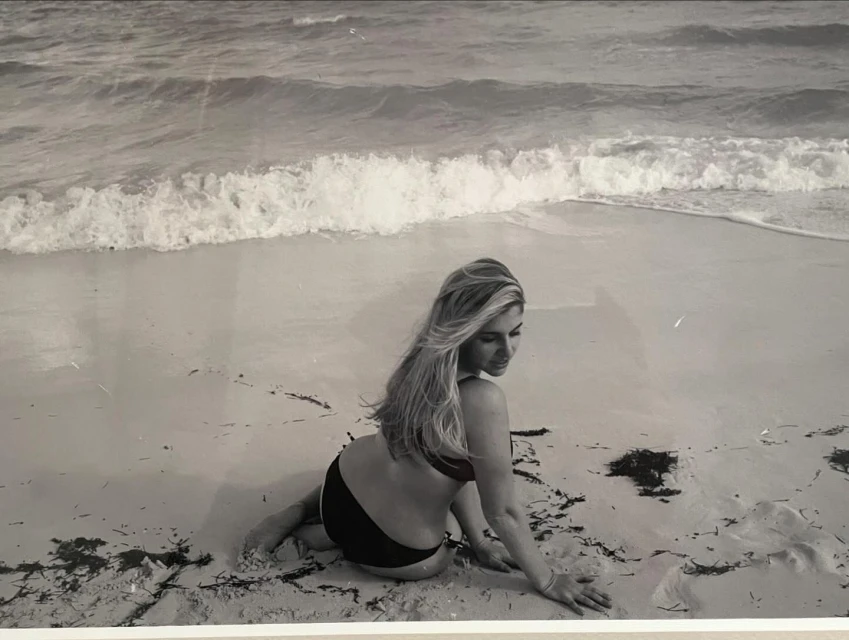 Monochrome photo of a woman in swimwear sitting on a sandy beach, with ocean waves in the background, captured in a vintage style.