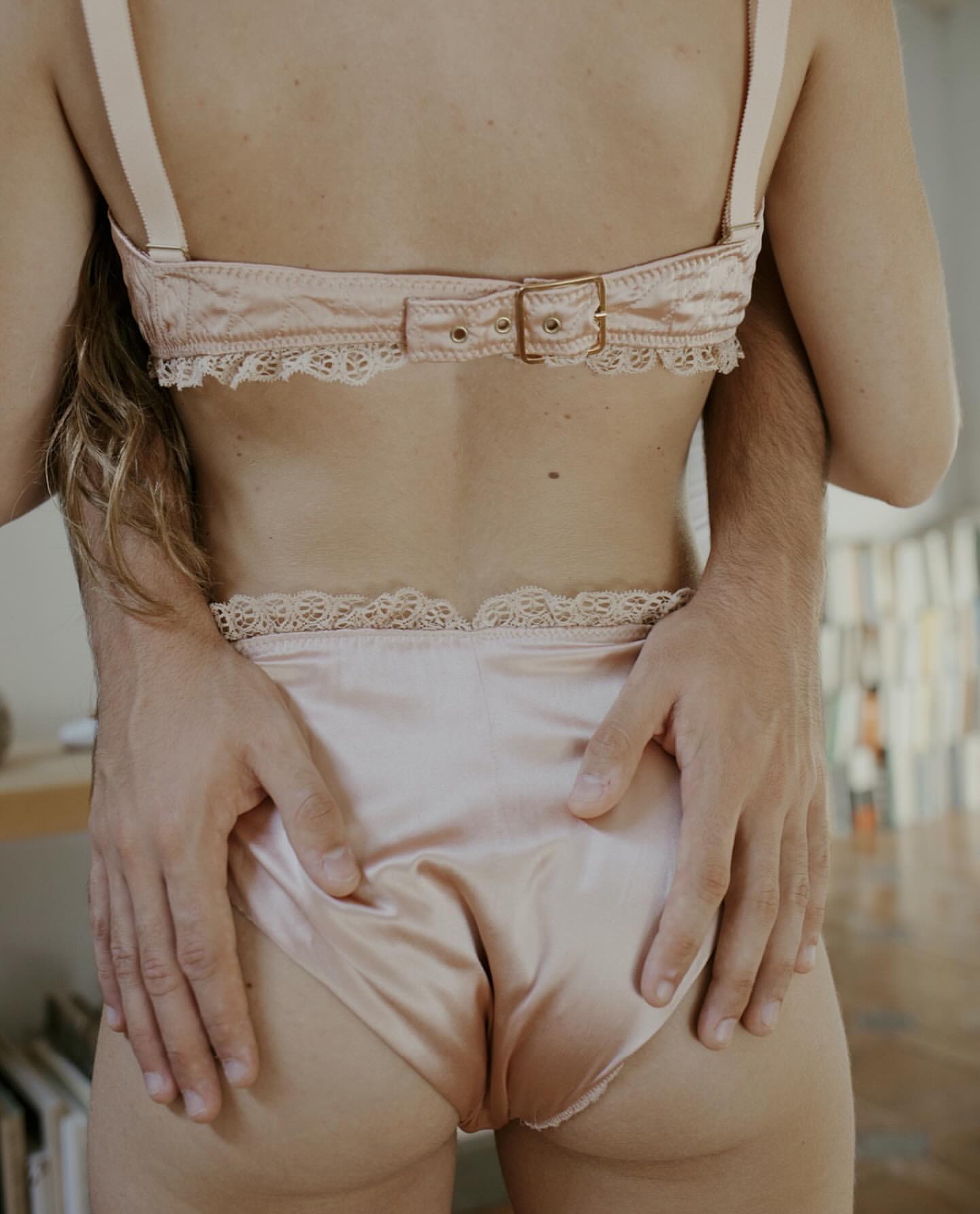 Woman poses in lingerie as a man's hands embrace her from behind inside a home with soft lighting, conveying intimacy and comfort.