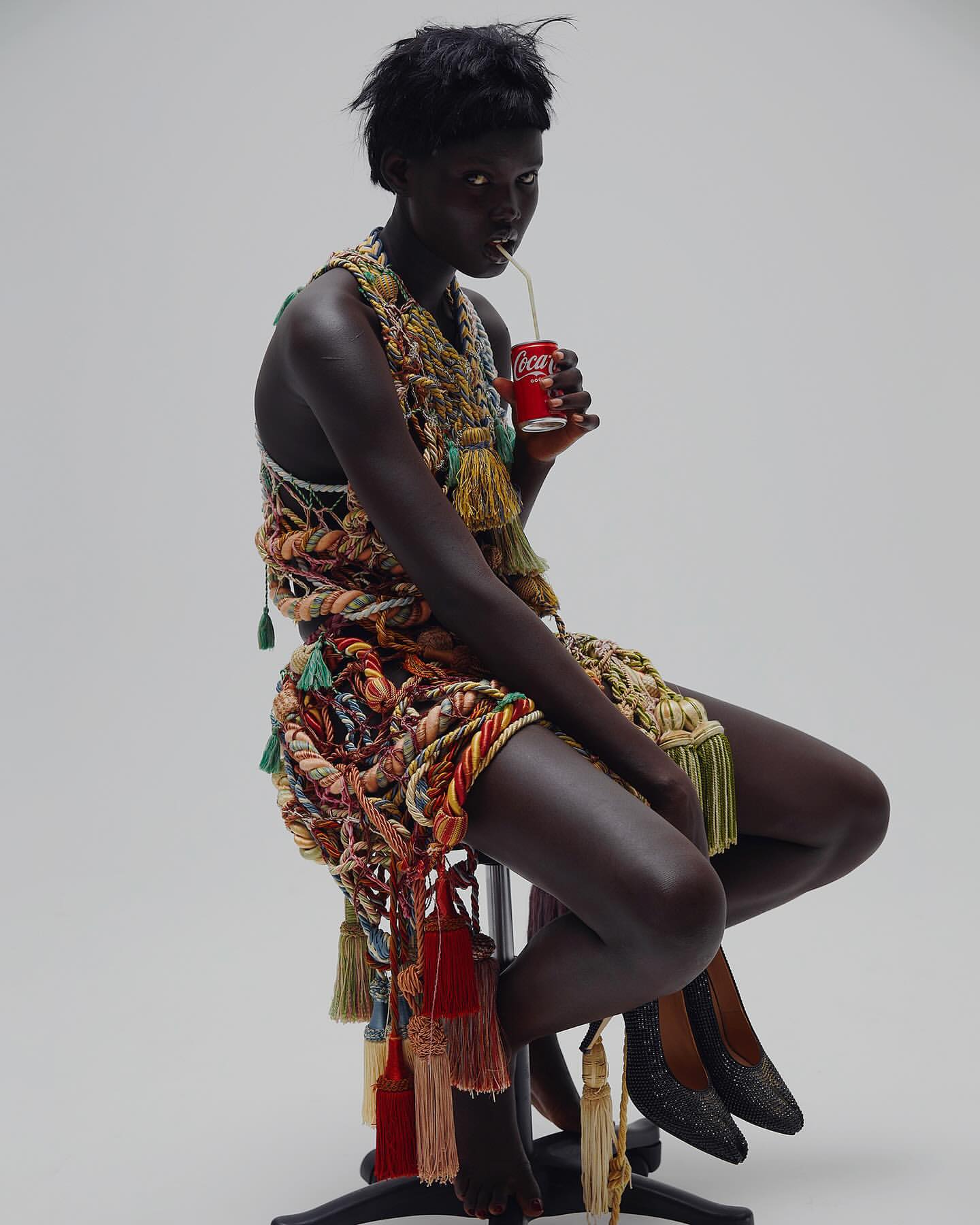Dark-skinned woman in rope dress sits, sips Coke with straw in studio. Fashion shot, white backdrop, high heels. One person posing.