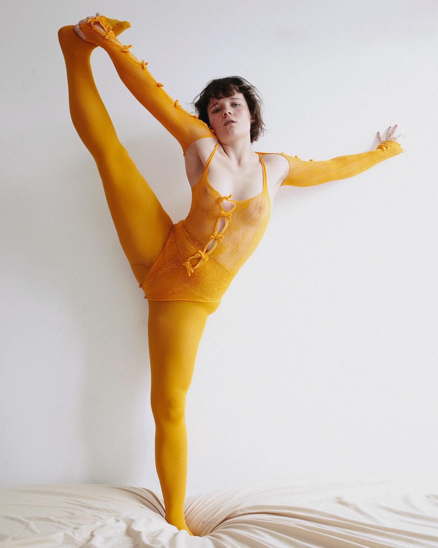 Woman in yellow bodysuit balances in unique yoga pose on bed against a plain white wall in a brightly lit indoor environment.