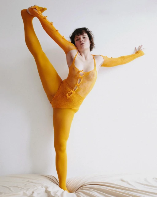 Woman in yellow bodysuit balances in unique yoga pose on bed against a plain white wall in a brightly lit indoor environment.