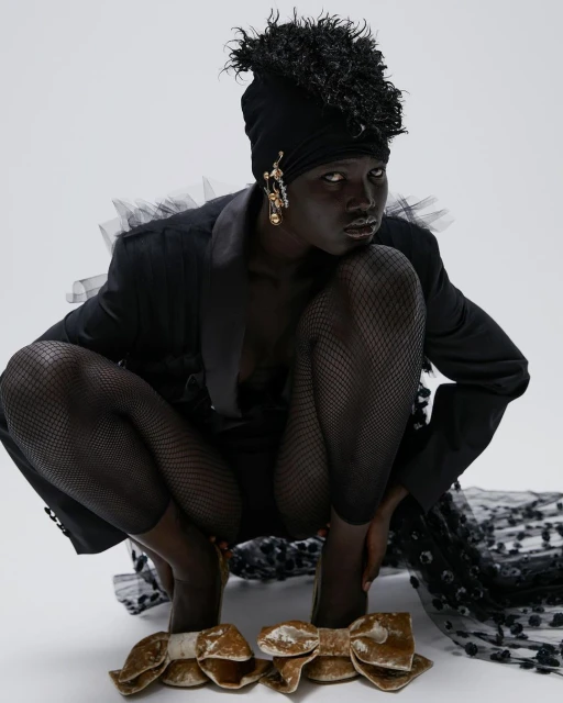 A fashion photo features a Black woman in a dark jacket, head wrap, and fishnet stockings, crouching in a white studio with oversized bow shoes.