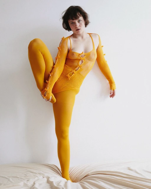 Woman in bright yellow bodysuit and tights standing on a bed with one leg raised, against a white wall in a studio setting.