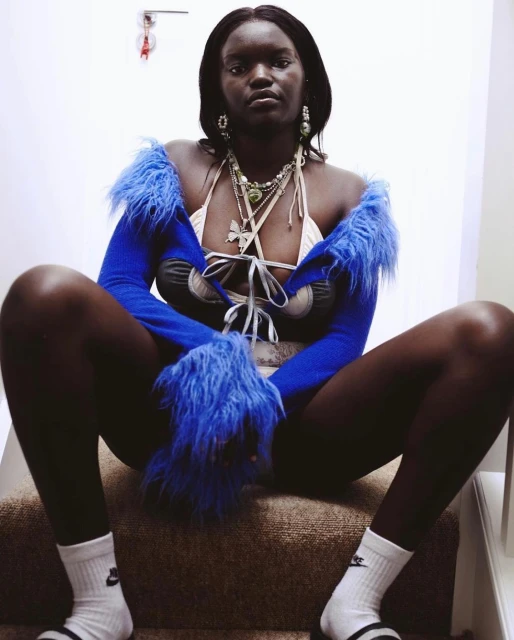 A dark-skinned woman sits on carpeted stairs, facing the camera with a neutral expression, wearing an open blue furry cardigan and jewelry.