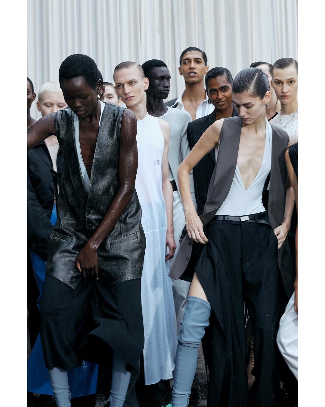 Runway models in vests and trousers backstage during a fashion show, featuring a diverse group of young adult men and women.