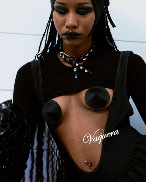 A young adult woman with dark eye makeup and dark lipstick, wearing a black top and bottom, looking down in a studio.