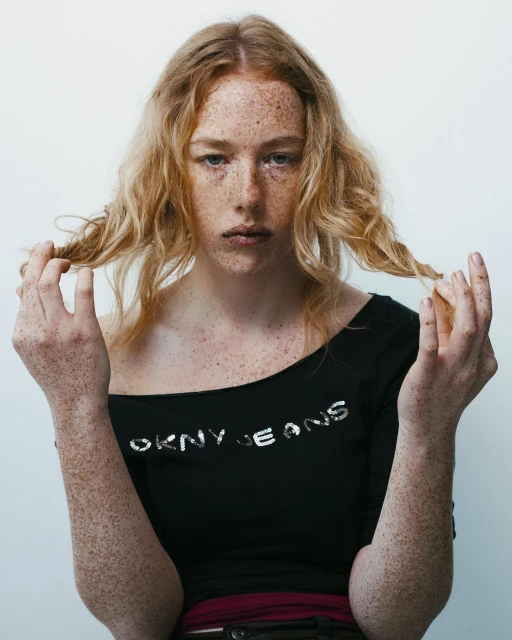 Portrait of a freckled woman with wavy light brown hair, wearing a black DKNY Jeans top, holding her hair in a photo studio setting.
