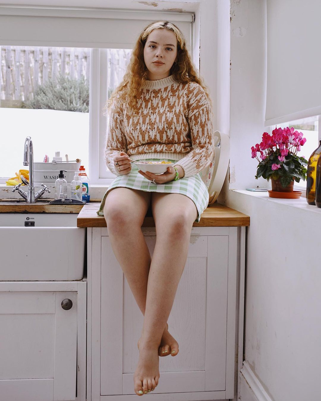 Young woman with auburn curly hair wearing a patterned sweater and skirt while sitting on a kitchen counter eating cereal.