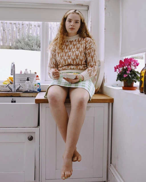 Young woman with auburn curly hair wearing a patterned sweater and skirt while sitting on a kitchen counter eating cereal.
