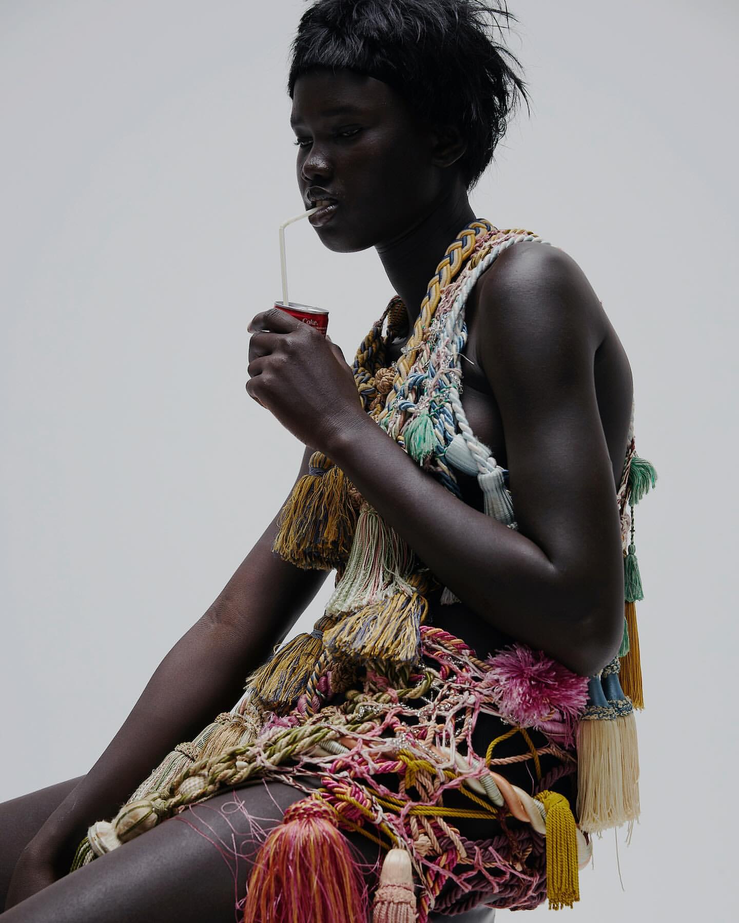 Woman with dark skin drinks from a can through a straw, wearing an elaborate tassel-embellished garment in a white studio setting.