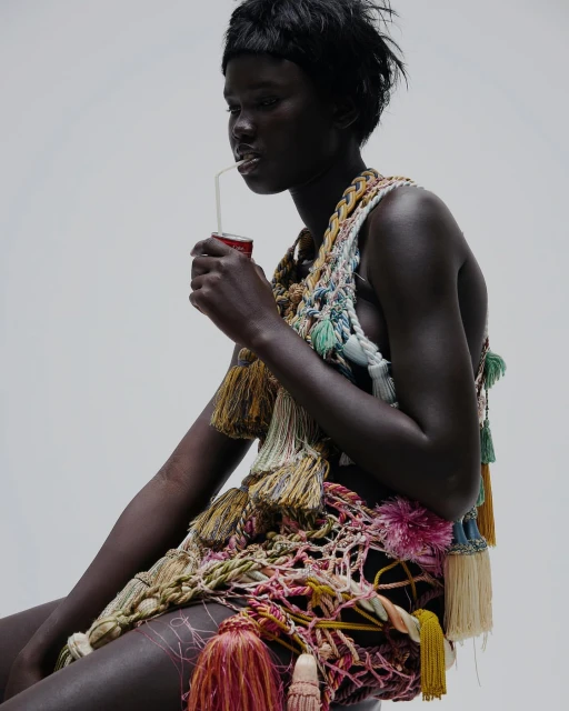 Woman with dark skin drinks from a can through a straw, wearing an elaborate tassel-embellished garment in a white studio setting.