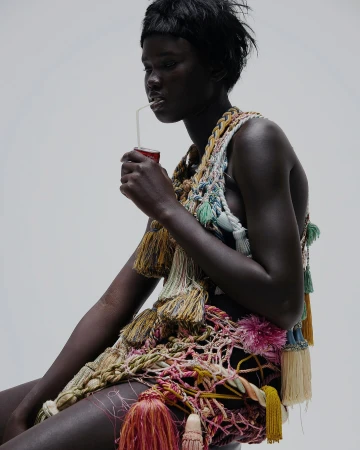 Woman with dark skin drinks from a can through a straw, wearing an elaborate tassel-embellished garment in a white studio setting.