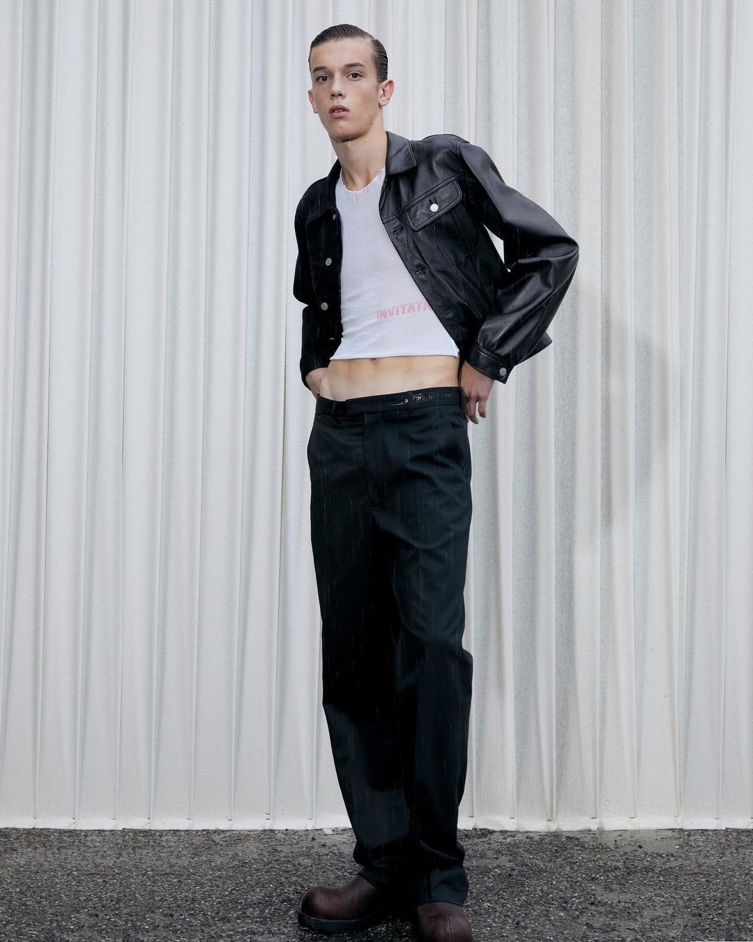 A slender male model poses in a white T-shirt, black leather jacket, and dark trousers against a textured white backdrop in a studio setting.