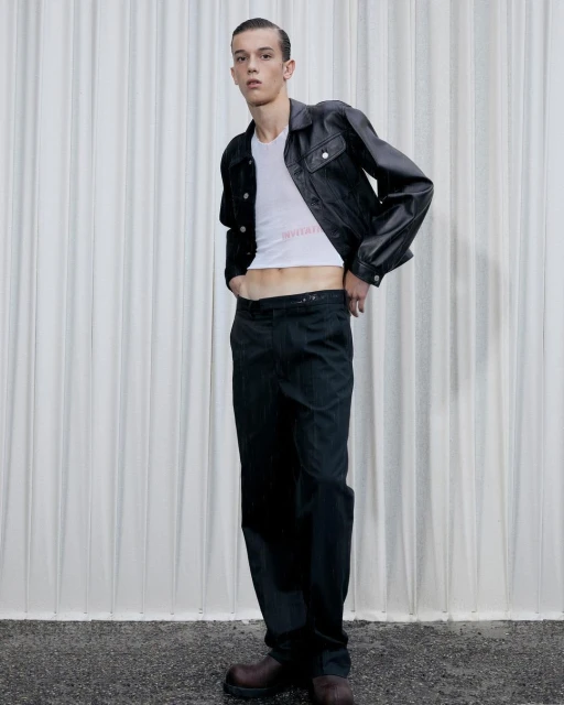 A slender male model poses in a white T-shirt, black leather jacket, and dark trousers against a textured white backdrop in a studio setting.