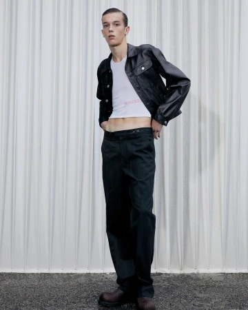 A slender male model poses in a white T-shirt, black leather jacket, and dark trousers against a textured white backdrop in a studio setting.
