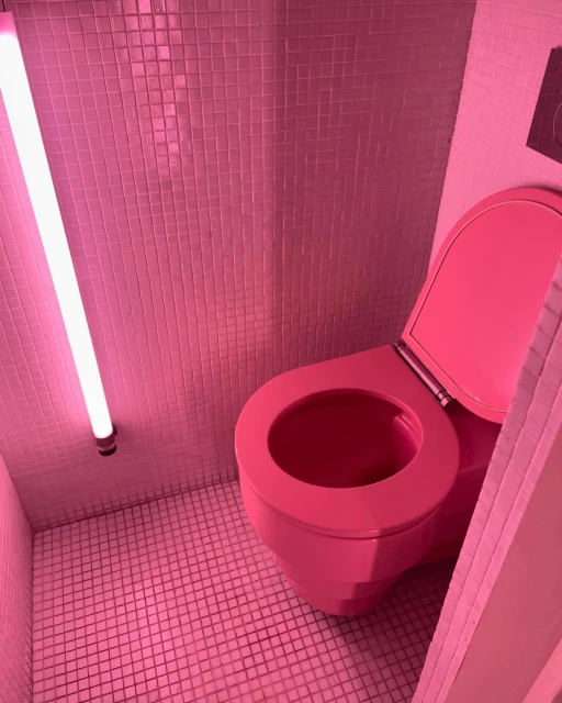 Bright pink restroom with a toilet, walls, and floor covered in small pink tiles. A pink neon light fixture illuminates the scene.
