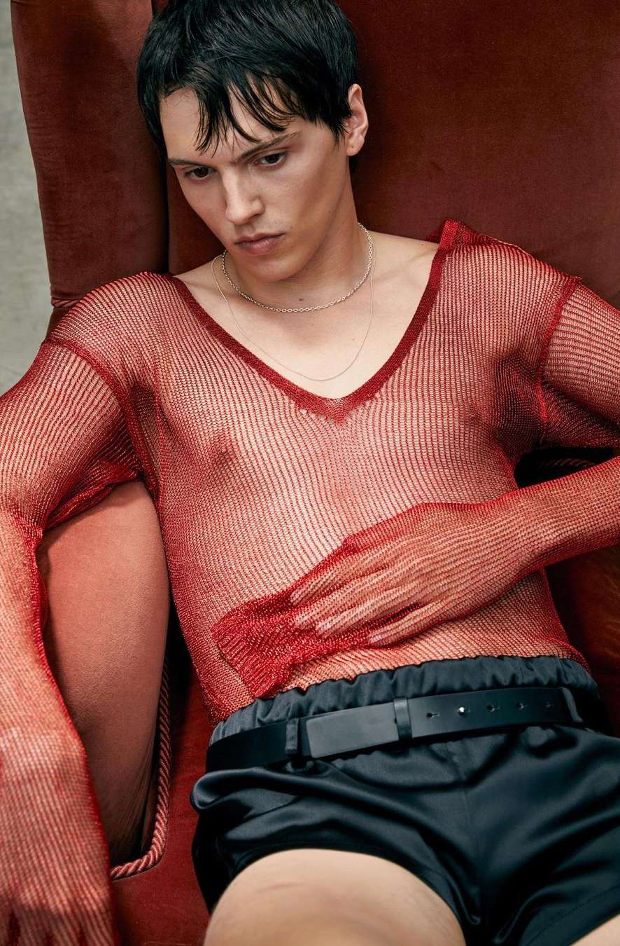 Male model poses indoors wearing a red mesh shirt and black shorts, sitting in a red chair against a neutral wall.