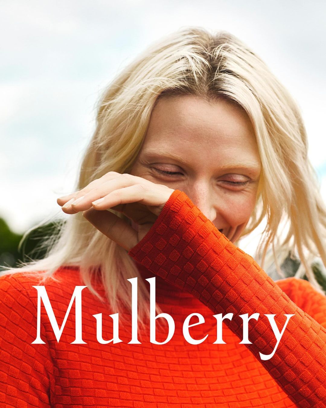 Close-up of a smiling woman with blonde hair wearing an orange sweater, with the text 'Mulberry' overlaid on the image.