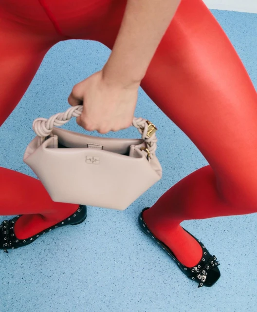 Close-up studio shot of a beige purse with knotted handle carried by a person wearing red tights and black flats with embellishments.