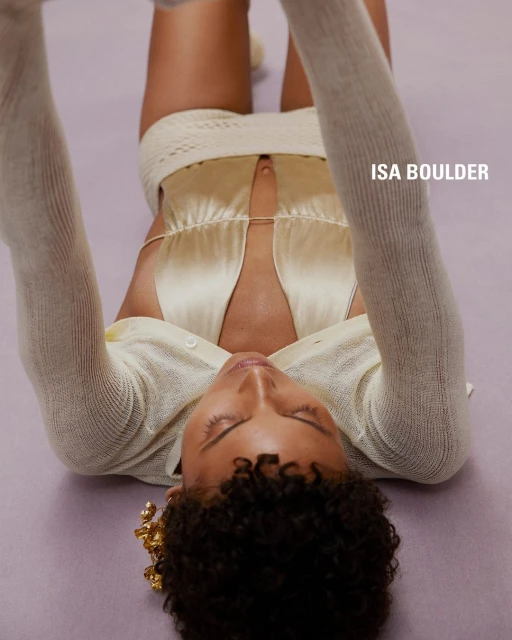 Woman in a cream-colored outfit laying on a soft, purple surface, holding her arms above her head in a studio shot, fashion-forward.