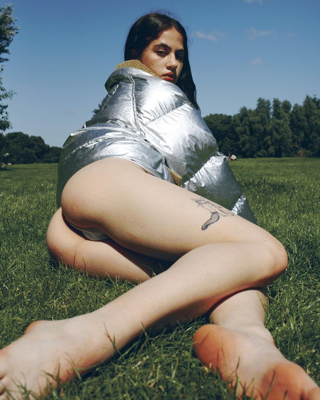 Brunette woman in a silver puffer jacket reclines on green grass under a blue sky, gazing at the camera with a serious expression.