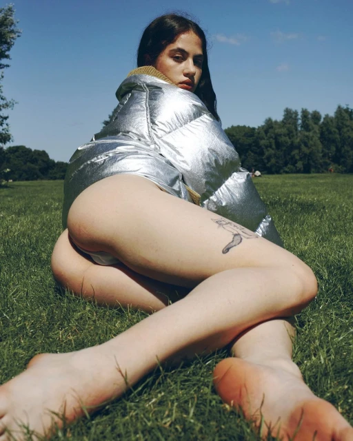 Brunette woman in a silver puffer jacket reclines on green grass under a blue sky, gazing at the camera with a serious expression.