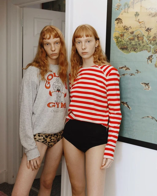 Two red-haired women in casual tops and underwear pose in an indoor setting with a vintage world map in the background.