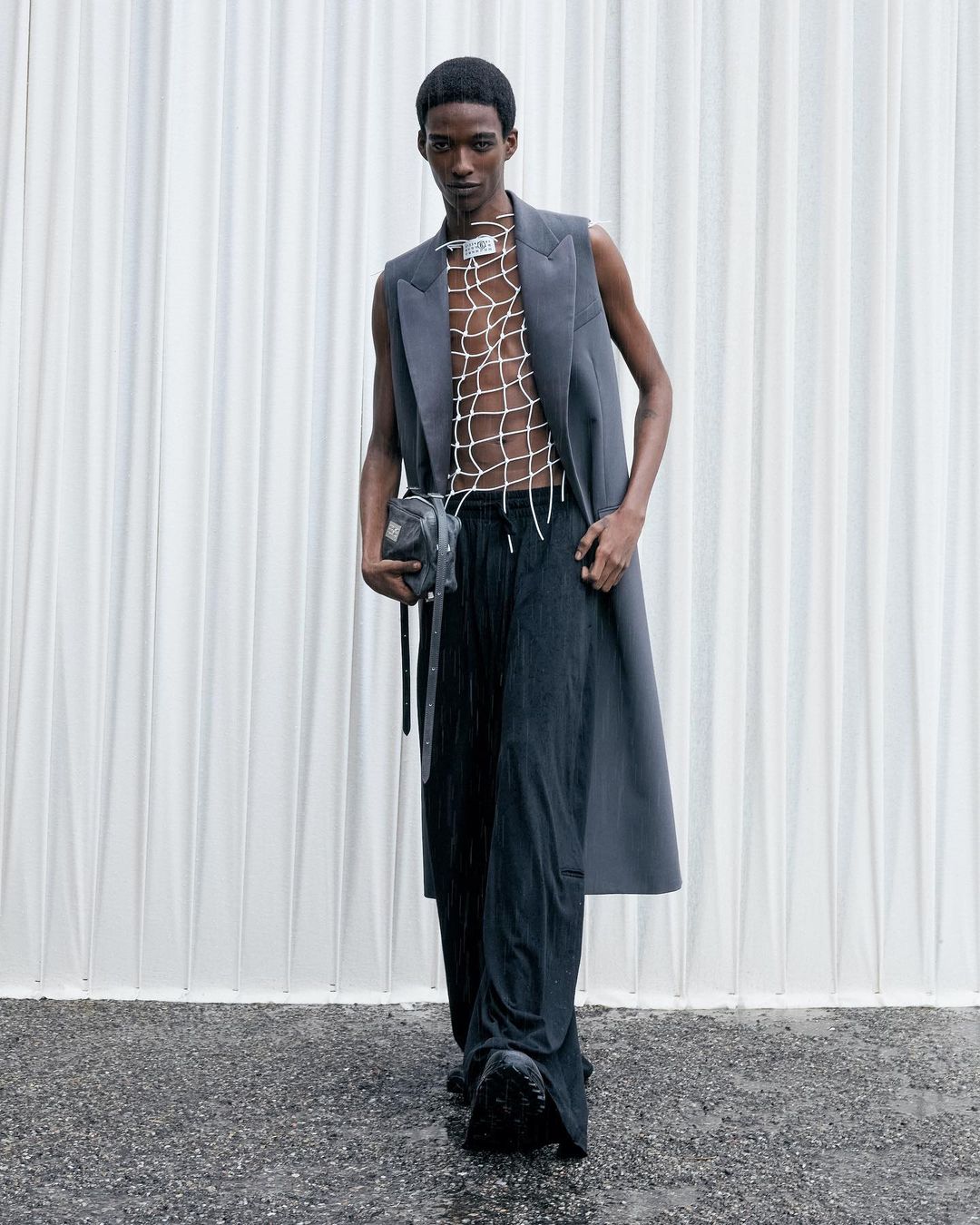 An adult male model poses in a sleeveless jacket, net top, wide-leg pants and bag against a bright, textured white background for a fashion shoot.