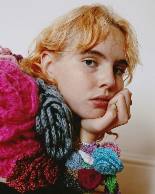 Portrait of a woman with freckles and curly red hair wearing a colourful floral sweater in a brightly lit interior space.