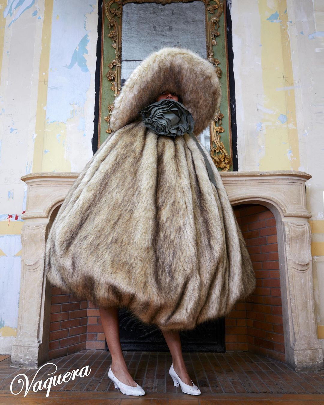 A model poses in a whimsical fur gown and large fur hat, standing in front of an ornate fireplace, and wearing white heeled shoes.