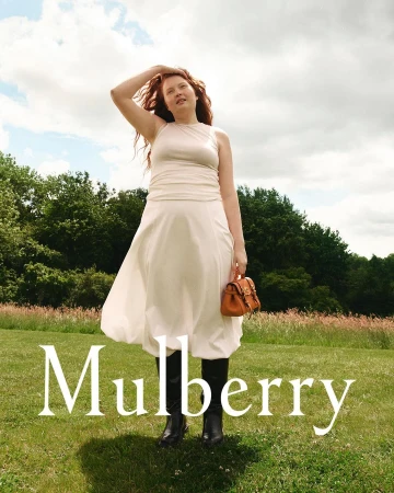 Red-haired model in an off-white dress holding a brown leather bag in a grassy park by Guen Fiore