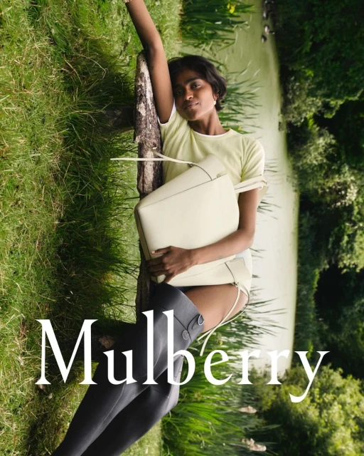 An East Indian woman poses with a Mulberry handbag outdoors by a pond, showcasing fashion in natural light against a backdrop of lush greenery.