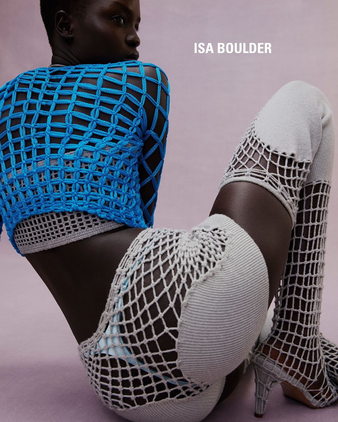 Fashion shot of a Black female model posing in a sitting position wearing a blue woven top and thigh-high knitted boots in a studio.