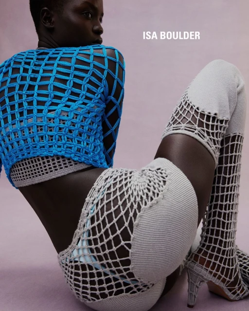 Fashion shot of a Black female model posing in a sitting position wearing a blue woven top and thigh-high knitted boots in a studio.