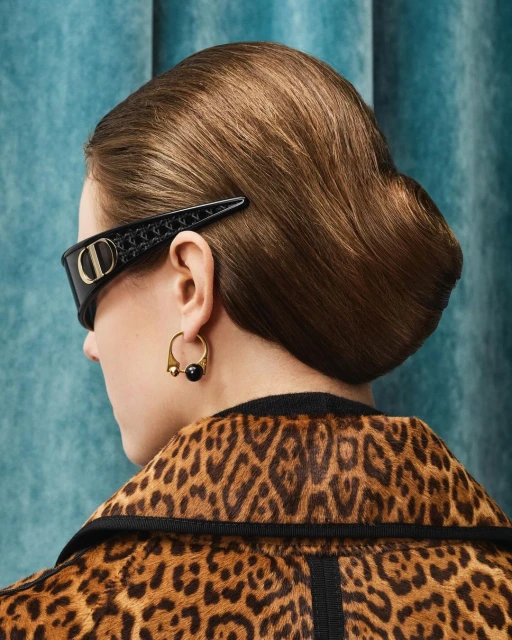 Woman with updo hairstyle wearing sunglasses with designer hair clip, gold hoop earrings, and animal print jacket against a blue backdrop.