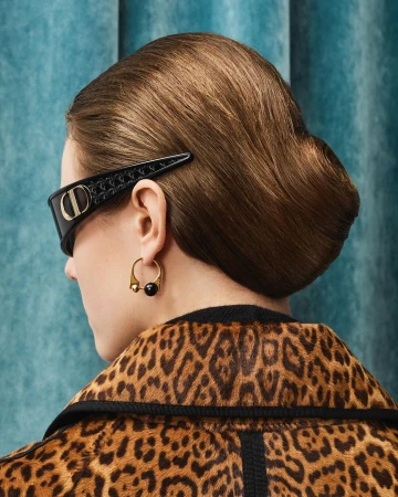Woman with updo hairstyle wearing sunglasses with designer hair clip, gold hoop earrings, and animal print jacket against a blue backdrop.