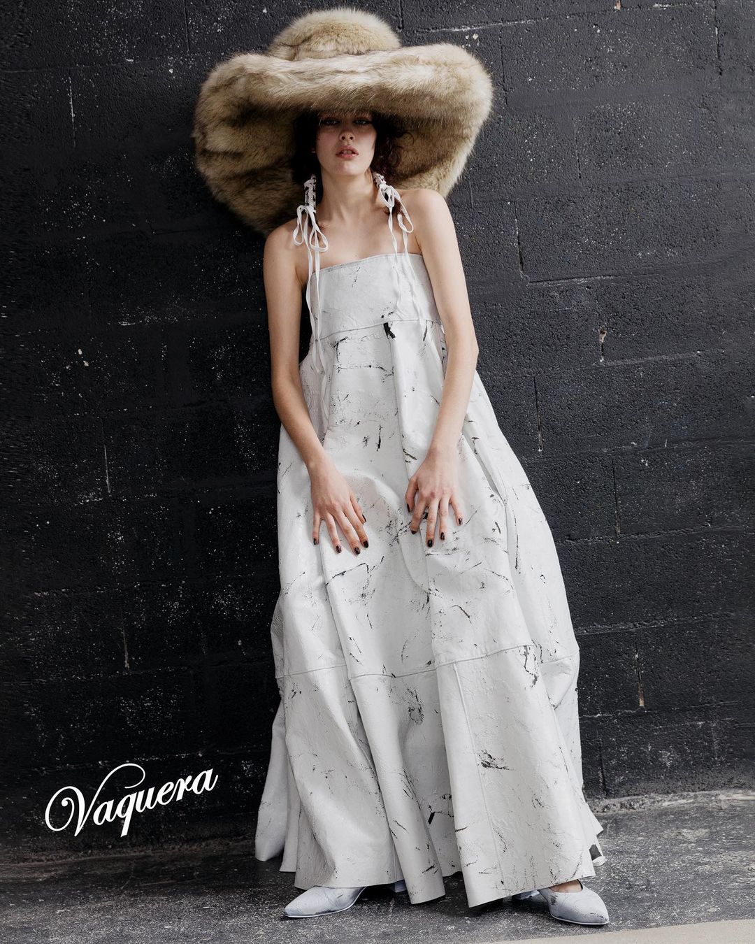 Woman in white printed dress and large fur hat posing in front of dark brick wall, showcasing fashion and style with neutral tones.