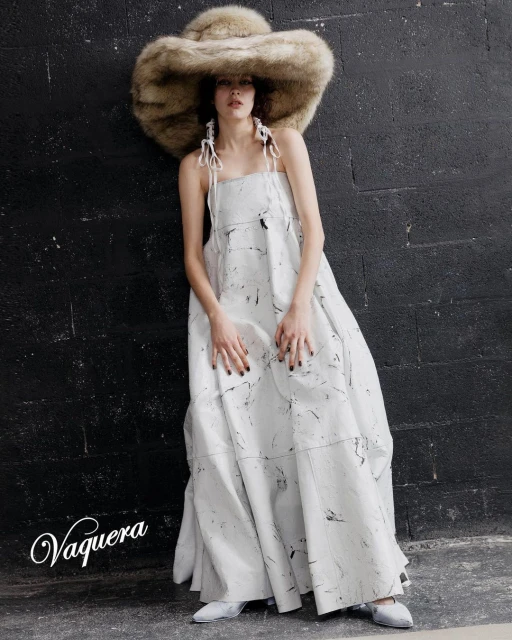 Woman in white printed dress and large fur hat posing in front of dark brick wall, showcasing fashion and style with neutral tones.