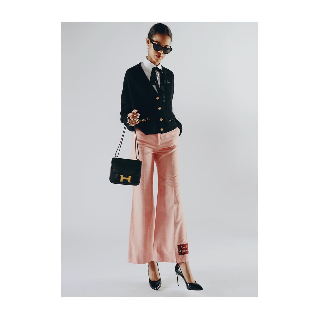 Fashionable woman in pink pants and a black cardigan poses with a purse against a bright backdrop, exuding elegance and style.