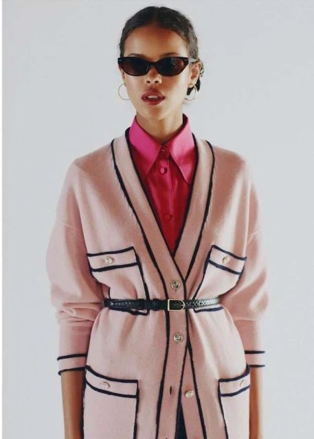 Woman in pink cardigan and sunglasses poses in a studio portrait, showcasing fashion and style against a clean white backdrop.