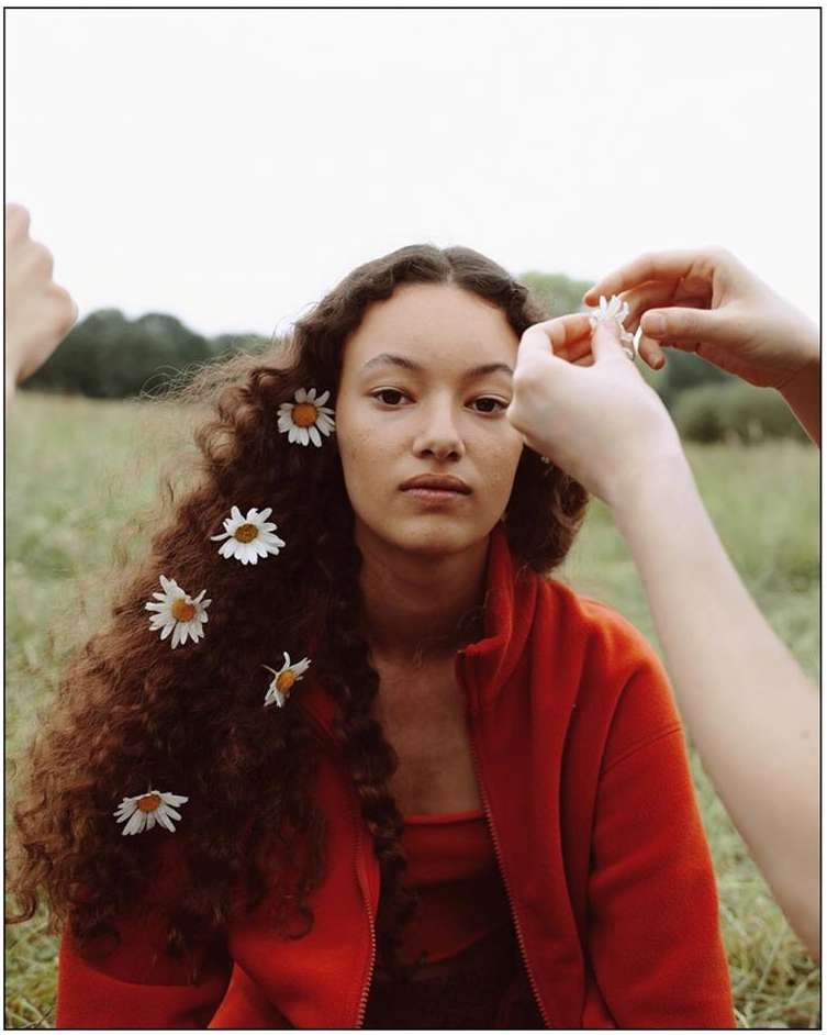 Young woman with daisies in her hair smiles serenely outdoors, wearing a red jacket, with hands arranging flowers in her hair.